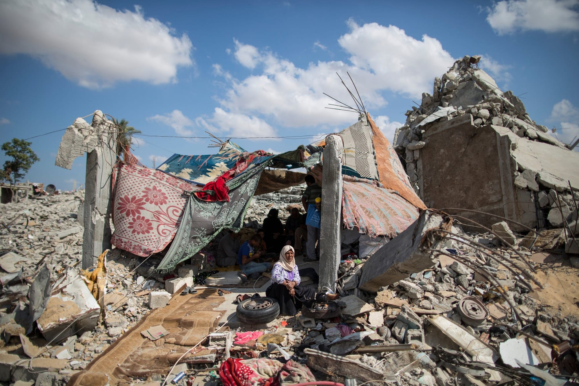 A woman in a headcarf crouches beofre a makeshift shelter of blankets and rugs, a tent built on a mound of rubble in the remains of a building in gaza.