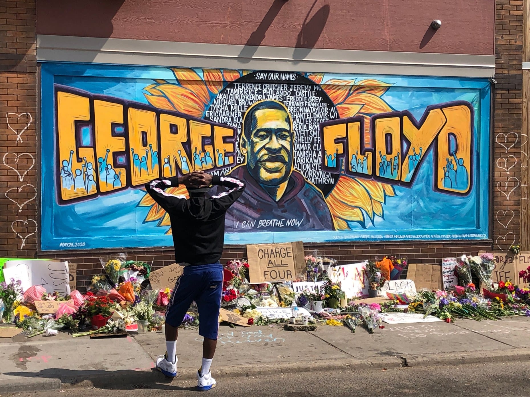 a young man stands, his hands on the back of his head, in front of the George Floyd mural in Minneapolis. A huge amount of flowers and cardboard protest signs lean against the brick wall beneath it