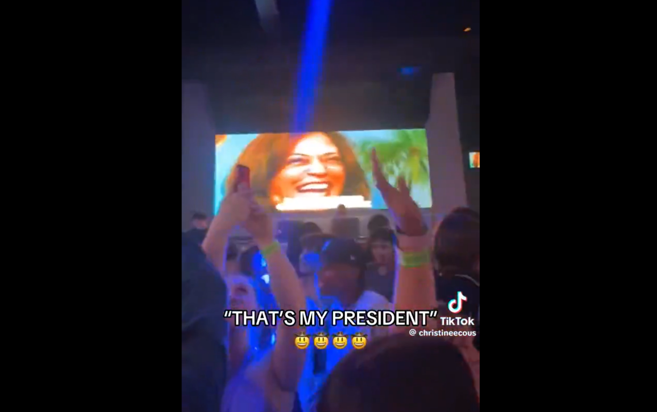 A club scene where people are dancing to a remix of Kamala laughing beneath her image projected on screens