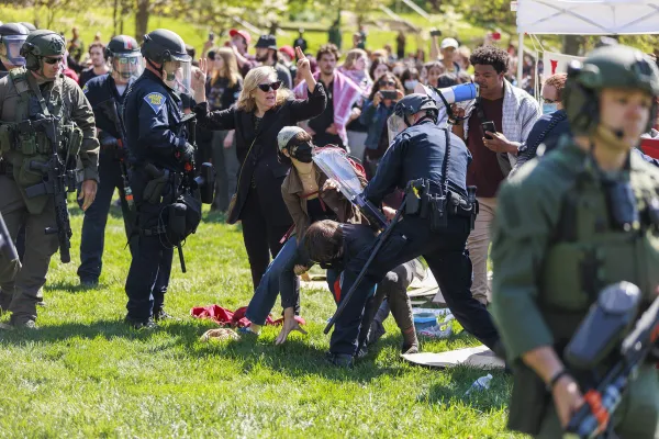 heavily militarized police frame a scene of a cop with a riot shield shoving two students to the ground o